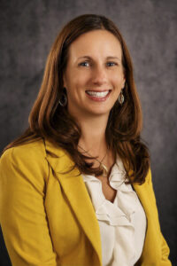 Deanna Schmitt, Interim Chair of the Dept. of Biological Sciences, wears a golden-yellow suit jacket over a white blouse, posing in front of a mottled gray background.