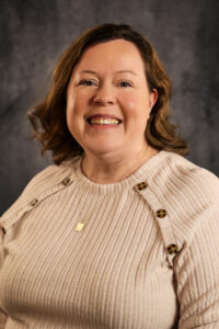 Bree Kibble, Administrative Services Coordinator for the College of Sciences is wearing a ribbed cream top with brown buttons at the shoulders, posing in front of a mottled gray background.