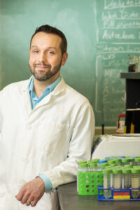 Dr. Joseph Horzempa, Interim Dean of the College of Sciences, in a white lab coat, leans against a lab bench holding lab equipment, that stands in front of a green chalkboard with lab notes written on it.