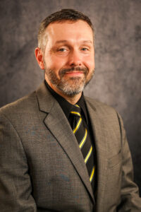 Dr. Jospeh Horzempa, Interim Dean of the College of Sciences, wearing a gray suit coat, black shirt and black and gold tie poses in front of a mottled gray background.